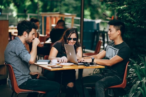 Three people at a cafe drinking coffee and looking over at a laptop, with books laid on the table