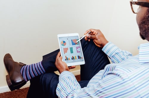 Over the shoulder perspective of a man sitting cross-legged and glancing at an iPad which shows statistics