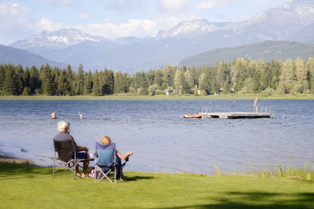 An old married couple sitting at the edge of a lake looking at trees and mountains in the distance